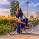 Photograph of a model in a long blue dress posing on a red bicycle in front of the Bosco Verticale buildings in Milan.